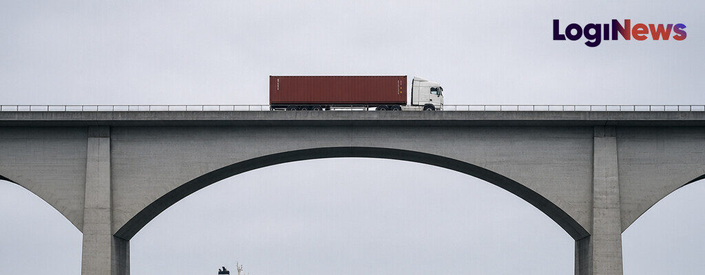 A truck carrying a container crosses a bridge while a container ship passes underneath